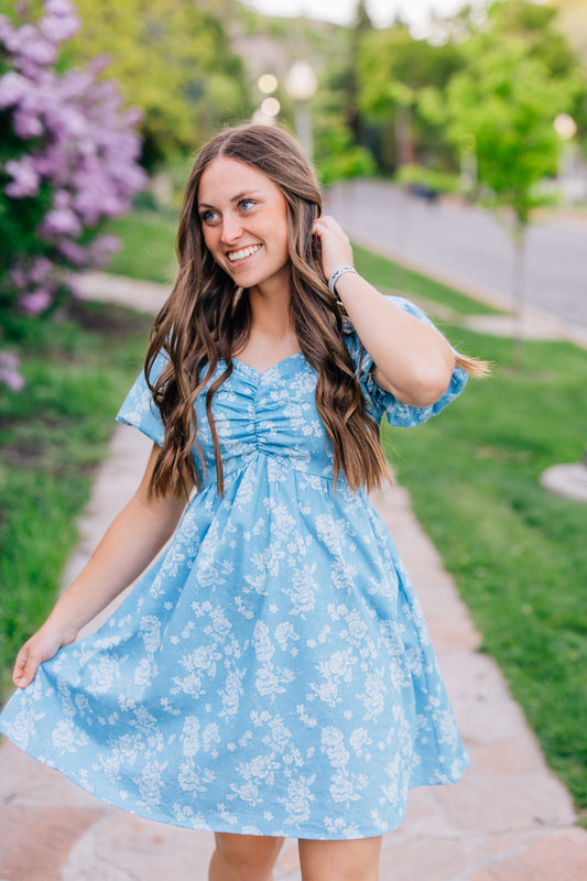 Woman in a blue floral dress standing outdoors with greenery in the background